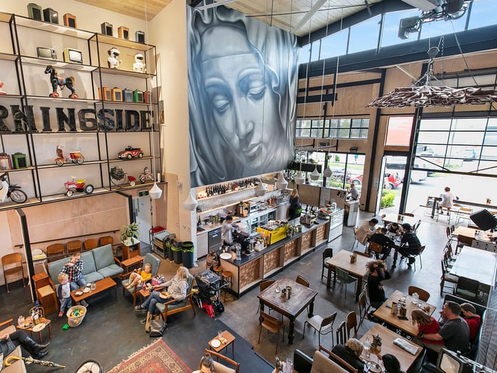 Bird's-eye view of double-height cafe with central bar, large face mural on wall, mixed seating throughout