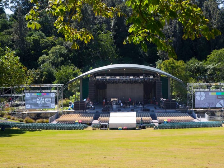 Outdoor amphitheatre with covered stage, seating, and large screens in parkland