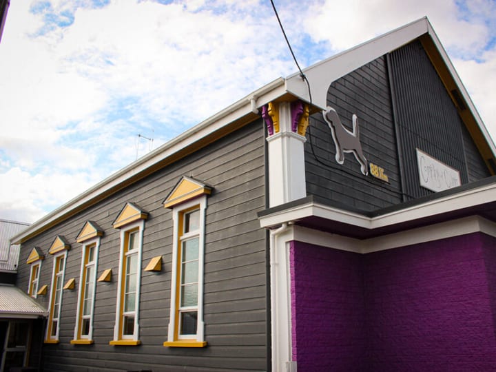 Grey weatherboard building with yellow-trimmed windows and purple brick lower section, street exterior