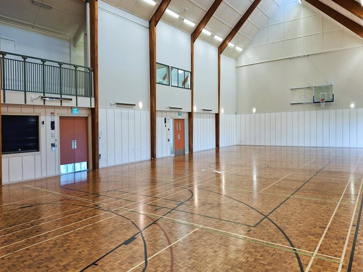 Sports hall with vaulted timber-beam ceiling, polished timber floor, mezzanine level, and basketball hoop