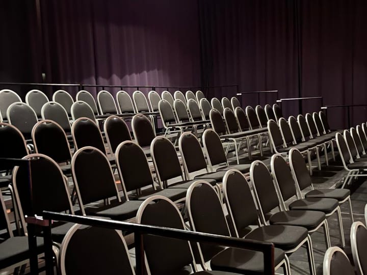 Dark theatre space with rows of black stackable chairs on a flat floor, dark curtain backdrop