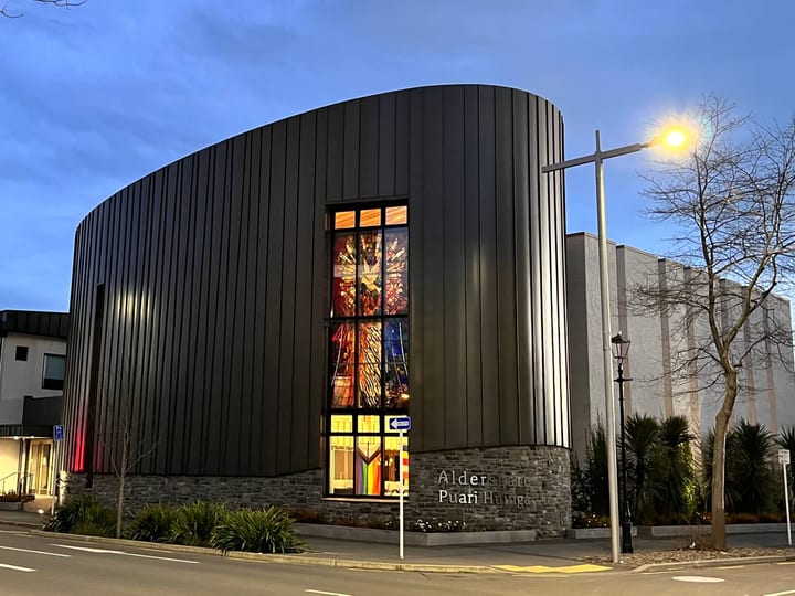Contemporary curved dark-clad church building at dusk with stained glass feature window