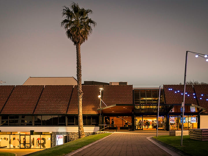 Low-rise arts centre exterior at dusk with a tall palm and festoon lights along the facade