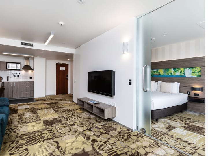 Open-plan hotel suite with kitchenette, frosted glass partition, and patterned carpet in green and gold tones