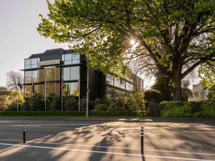 Dark multi-storey building with vertical timber screens, large trees framing the street frontage