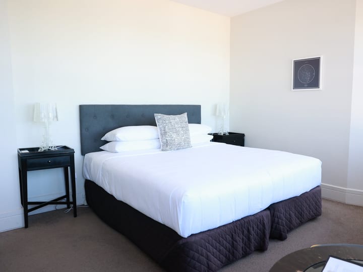 Hotel room with upholstered king bed, grey headboard, white linen, side tables and framed print