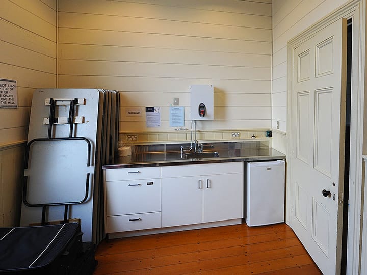 Small kitchenette with stainless bench, white cabinetry, and folded tables leaning against wall