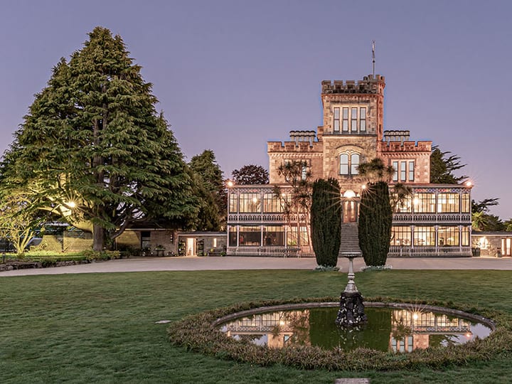 A lit stone castle with a tower and glasshouse wing at dusk, framed by tall trees and a formal lawn with a circular fountain pool
