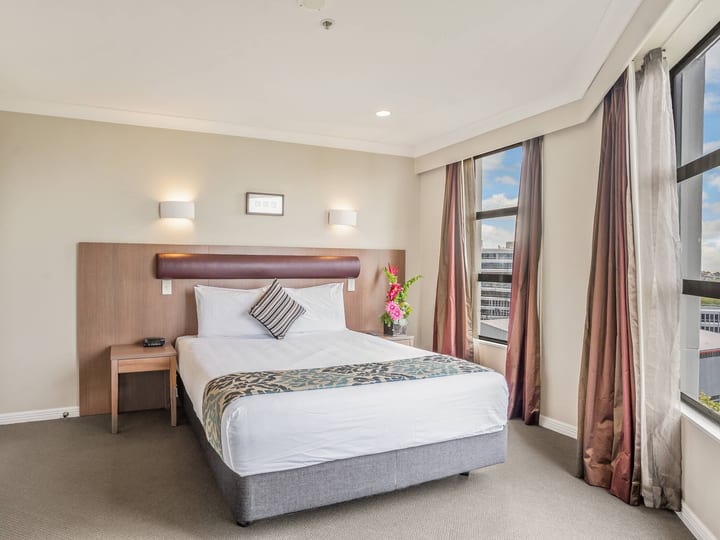 Hotel bedroom with timber-panel headboard, double bed and large windows overlooking rooftops