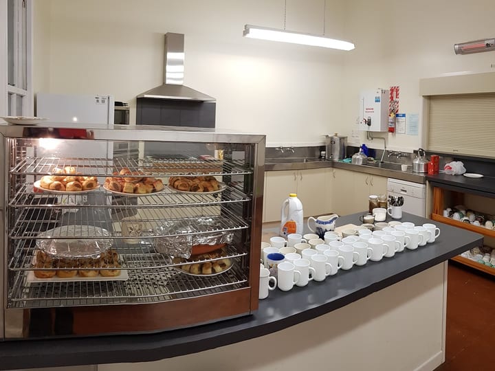 Hall kitchen with stainless benches, display warmer with baked goods, and mugs lined up on counter