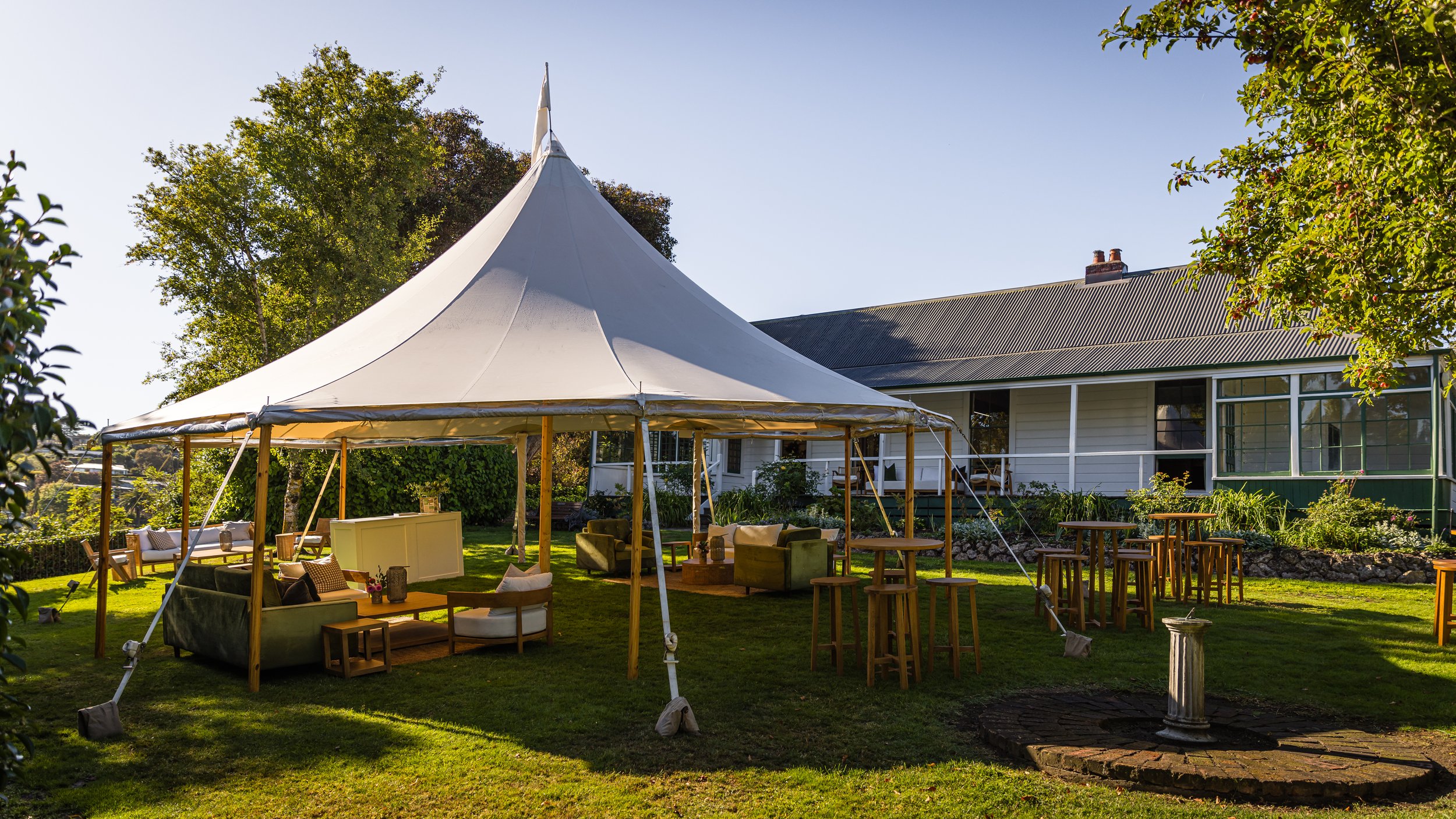 White marquee on a lawn with lounge seating and tall tables beside a single-storey house