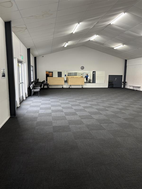 Large empty hall with grey carpet tiles, mirrors on the far wall and stacked chairs