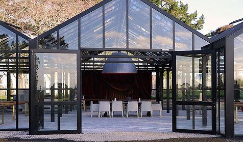Black steel glasshouse interior with white chairs and dark hanging pendant, surrounded by trees