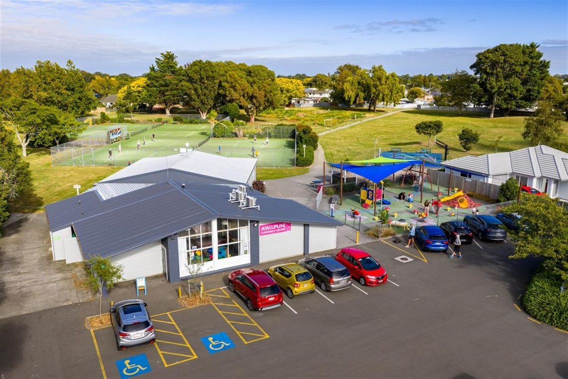 Aerial view of a community centre, car park, playground and tennis courts on green reserve