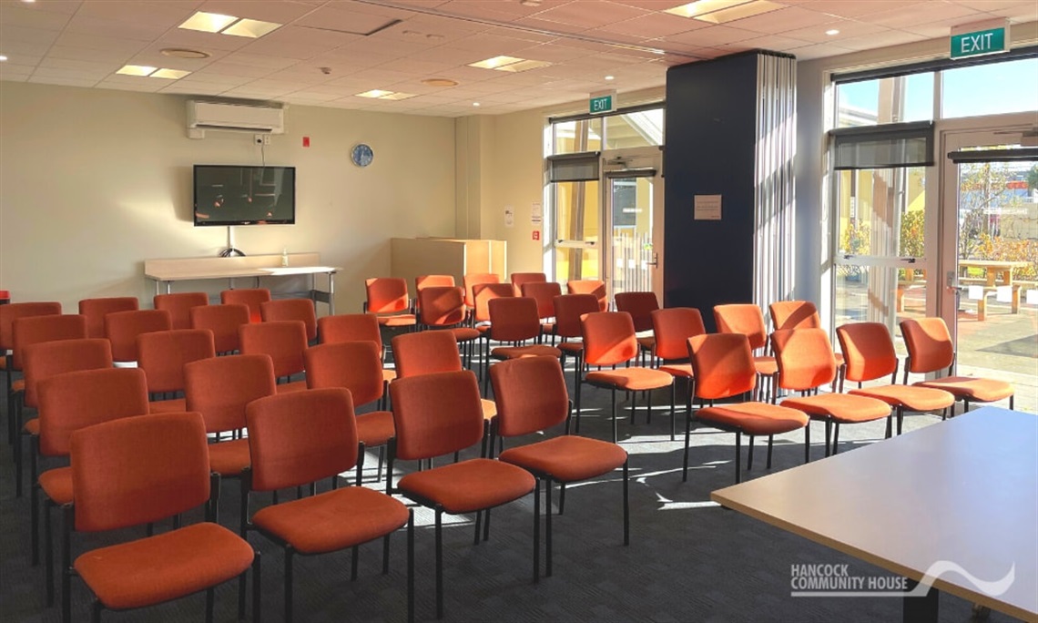 Meeting room with rows of orange chairs facing a wall-mounted screen